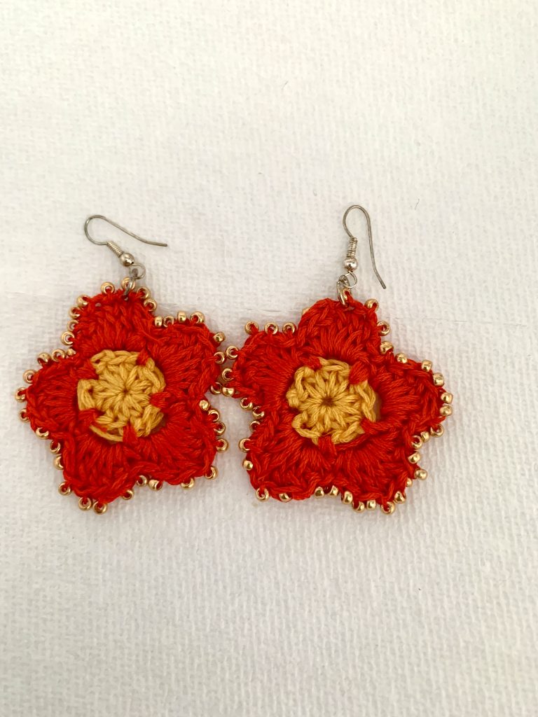 Close-up photograph of a handcrafted crochet red flower with five petals, featuring a crocheted yellow center. The edges of the red petals are adorned with golden-colored seed beads. The flower is attached to a French ear wire hook