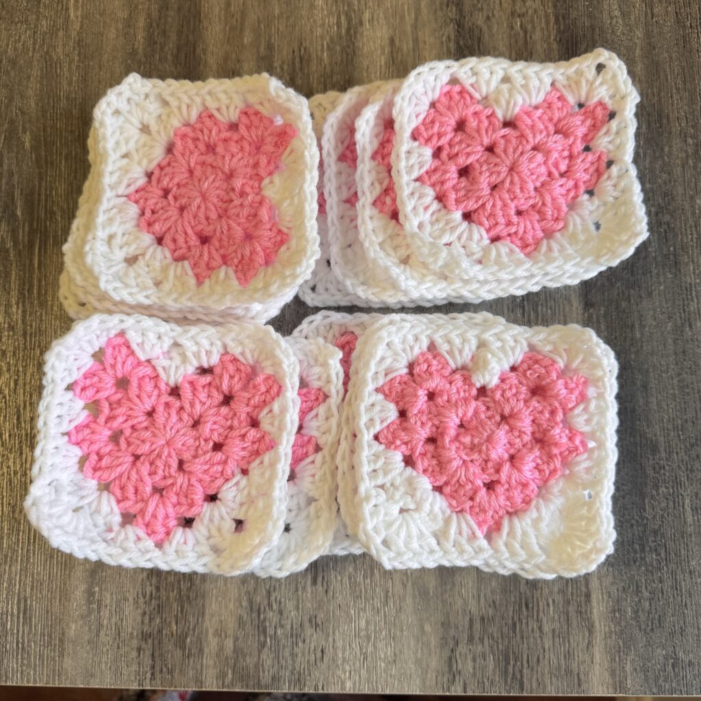 A collection of pink heart granny squares crocheted by Crochetsmash, neatly stacked on a wooden surface. Each square features a pink heart in the center with a white border, showcasing intricate craftsmanship as part of a crochet tutorial project.